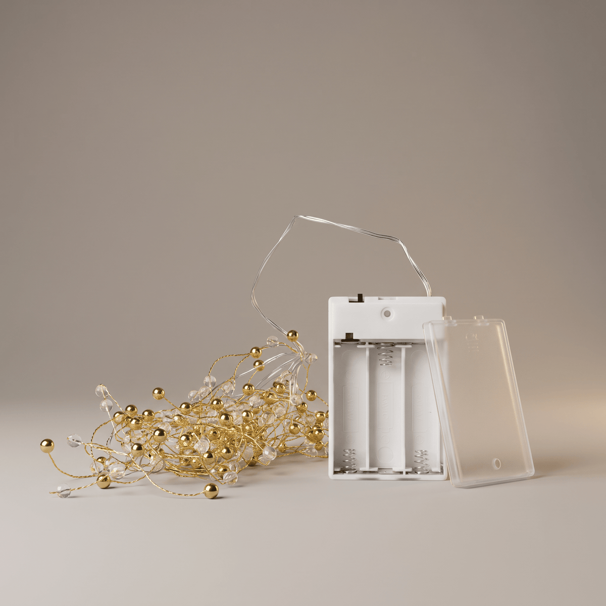 A set of gold LED fairy string lights with clear glass beads, shown next to a white battery compartment holder with its transparent plastic cover off, on a neutral beige background.