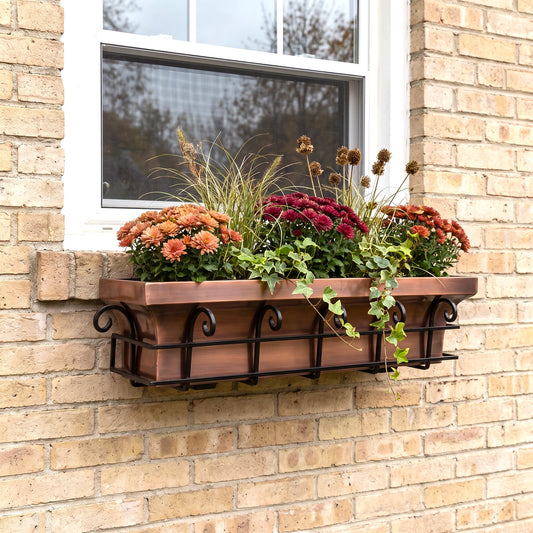 h potter brand copper-colored metal planter box with a black scrollwork base, mounted on a beige brick wall beneath a white window. The planter is filled with orange, red, and burgundy mums, ornamental grass, and trailing ivy.
