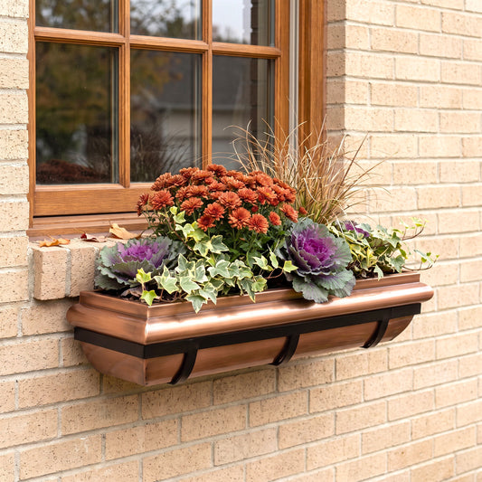 A copper-colored metal window box planter mounted on a beige brick wall beneath a wooden window, filled with orange flowers, purple cabbage, and green foliage.