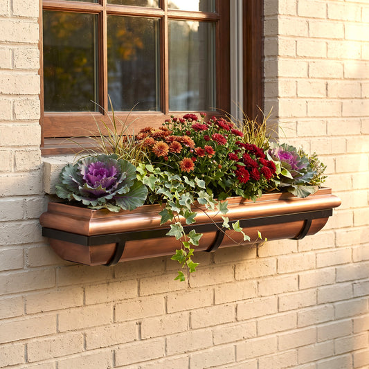 A copper-colored metal window box planter with black brackets, mounted on a beige brick wall beneath a wooden window and filled with colorful fall flowers and green foliage.