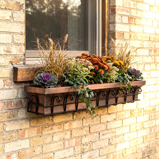 h potter brand rectangular copper-toned metal planter box with decorative scrollwork, mounted on a brick wall beneath a window and filled with ornamental grasses, succulents, and orange flowers.