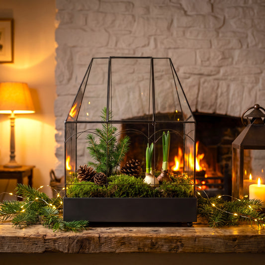 h potter glass terrarium with a house-shaped frame, filled with green moss, pine branches, pinecones, and small green shoots, displayed on a rustic wooden mantel in front of a cozy fireplace with a white brick wall.