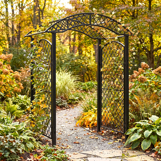 H Potter black metal garden arbor arch with lattice trellis panels stands open on a gravel path surrounded by autumn foliage and plants in a garden setting.