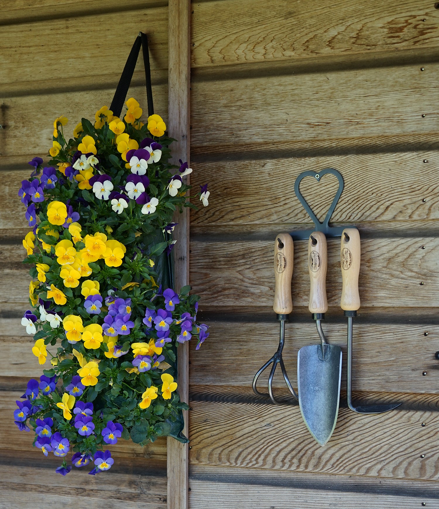 H Potter × DeWit garden tools displayed on heart-shaped metal hanger on garden wall with flowers