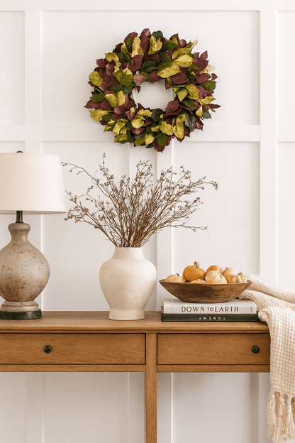 decorative wreath with red green and gold leaves on a white wall above a desk with white vase