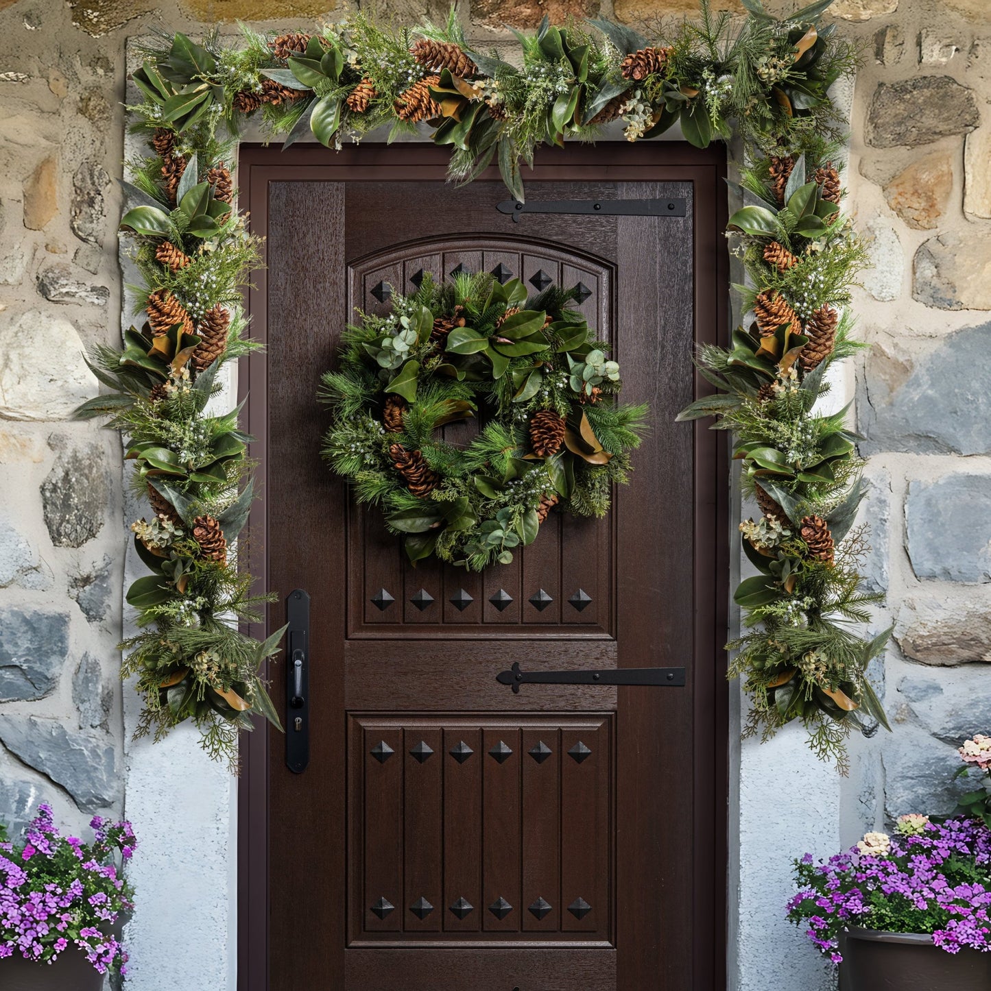 Decorative wreath on a brown door with greenery and pinecones, flanked by stone wall and potted plants.