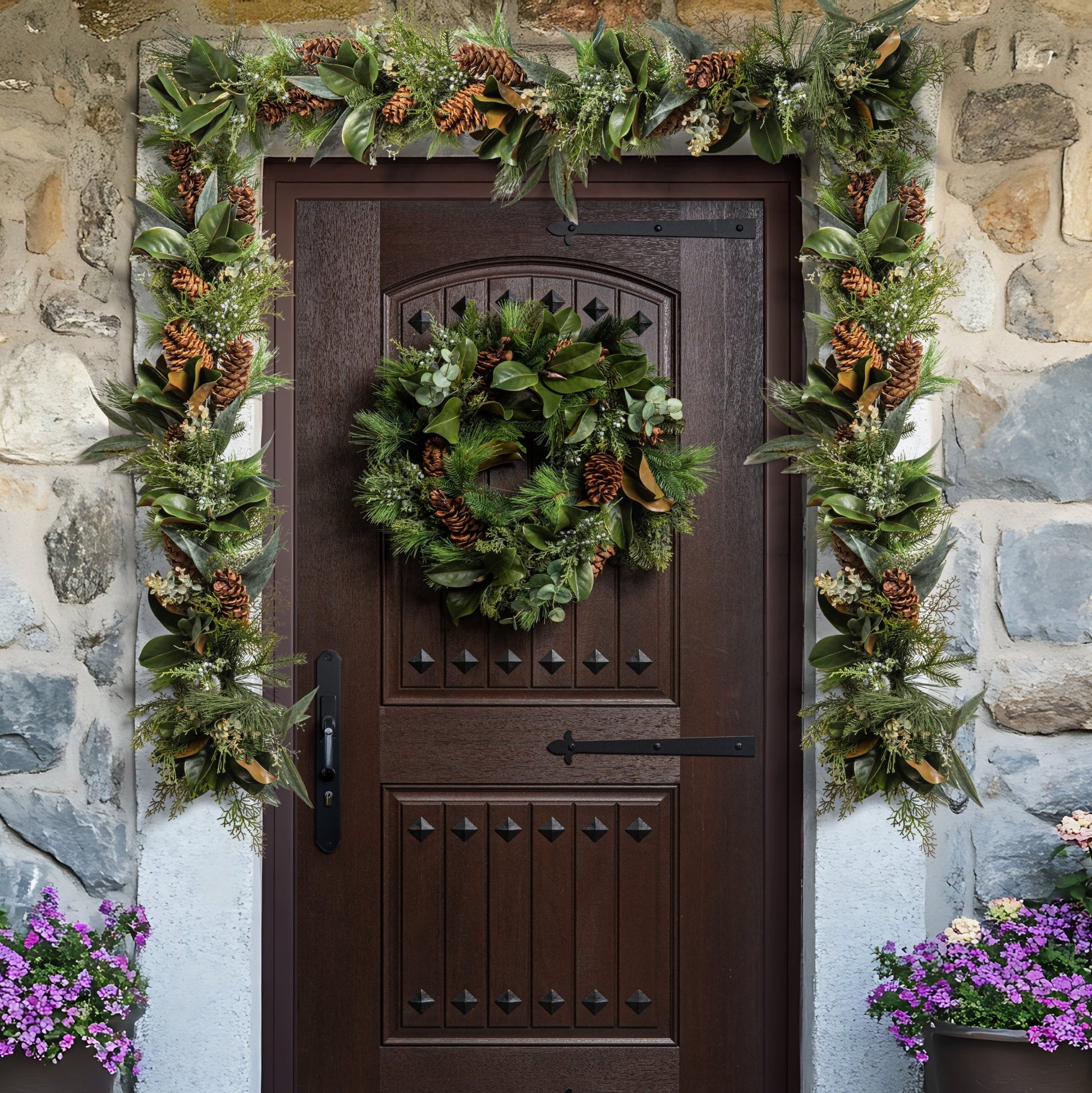 Decorative wreath on a brown door with greenery and pinecones, flanked by stone wall and potted plants.
