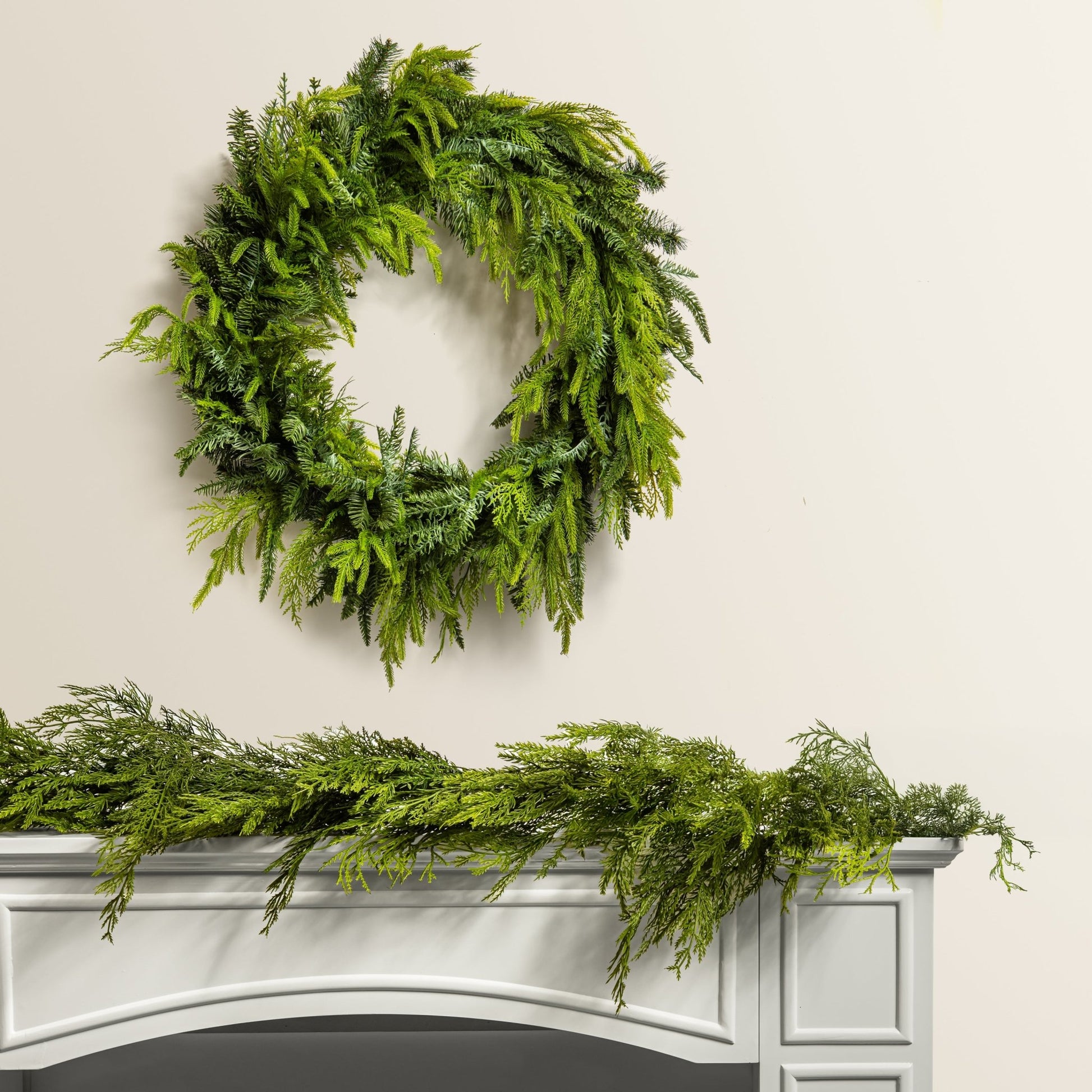 Green christmas wreath and garland on a white fireplace mantel against a beige wall.