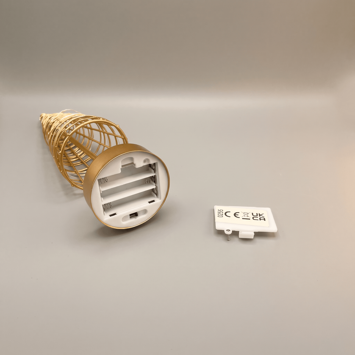 A gold wire tree with its white battery compartment visible and a small white CE mark card placed beside it on a neutral gray background.