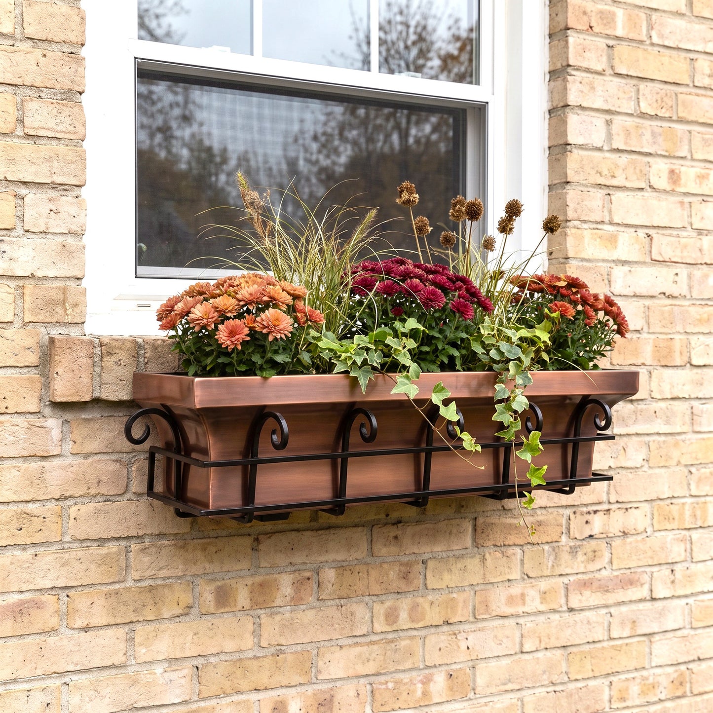 h potter brand copper-colored metal planter box with a black scrollwork base, mounted on a beige brick wall beneath a white window. The planter is filled with orange, red, and burgundy mums, ornamental grass, and trailing ivy.