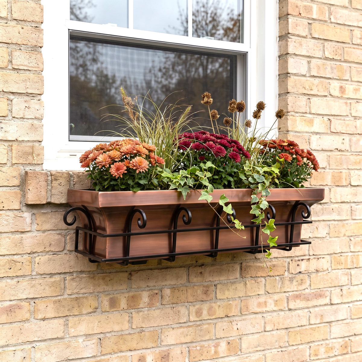 h potter brand copper-colored metal planter box with a black scrollwork base, mounted on a beige brick wall beneath a white window. The planter is filled with orange, red, and burgundy mums, ornamental grass, and trailing ivy.