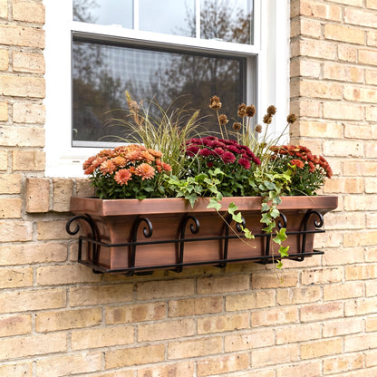 h potter brand copper-colored metal planter box with a black scrollwork base, mounted on a beige brick wall beneath a white window. The planter is filled with orange, red, and burgundy mums, ornamental grass, and trailing ivy.