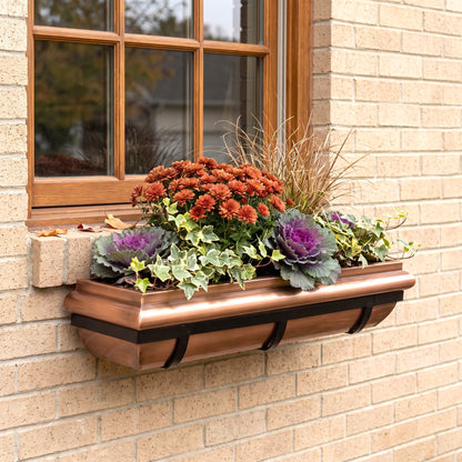 A copper-colored metal window box planter mounted on a beige brick wall beneath a wooden window, filled with orange flowers, purple cabbage, and green foliage.