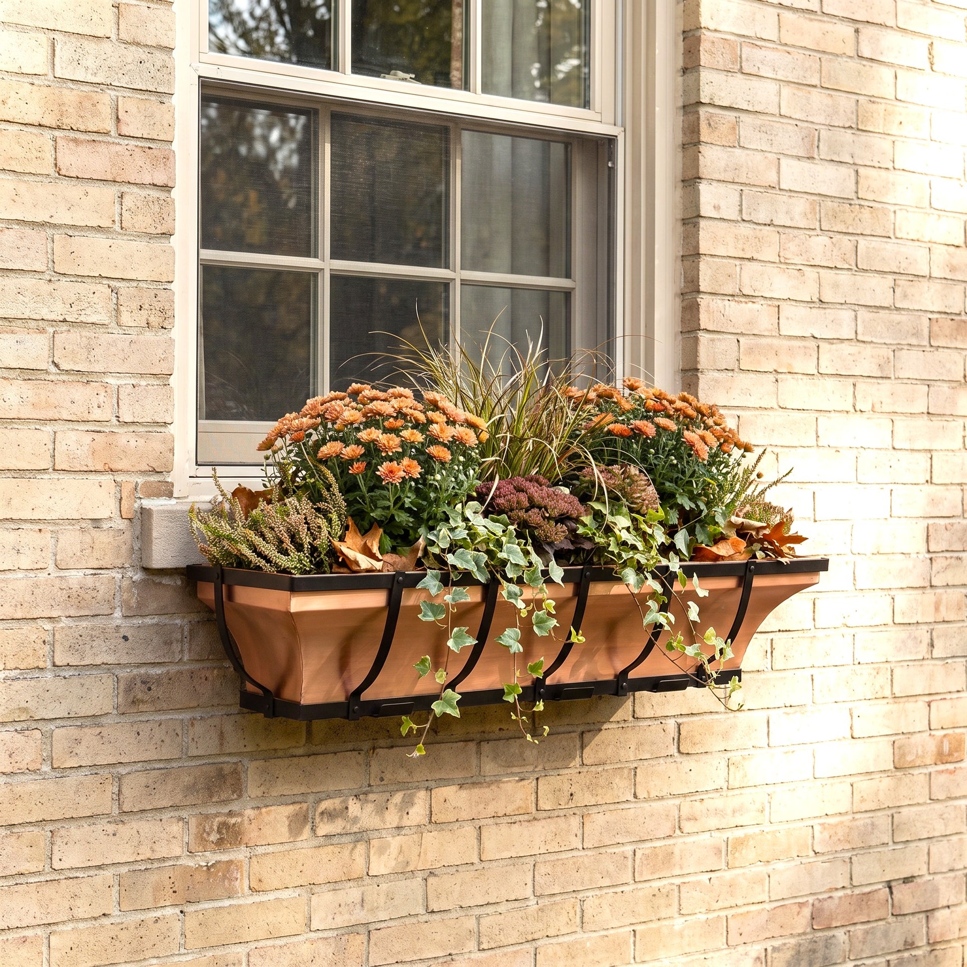 h potter brand window box with black metal brackets mounted on a beige brick wall beneath a window, filled with orange flowers and green foliage.