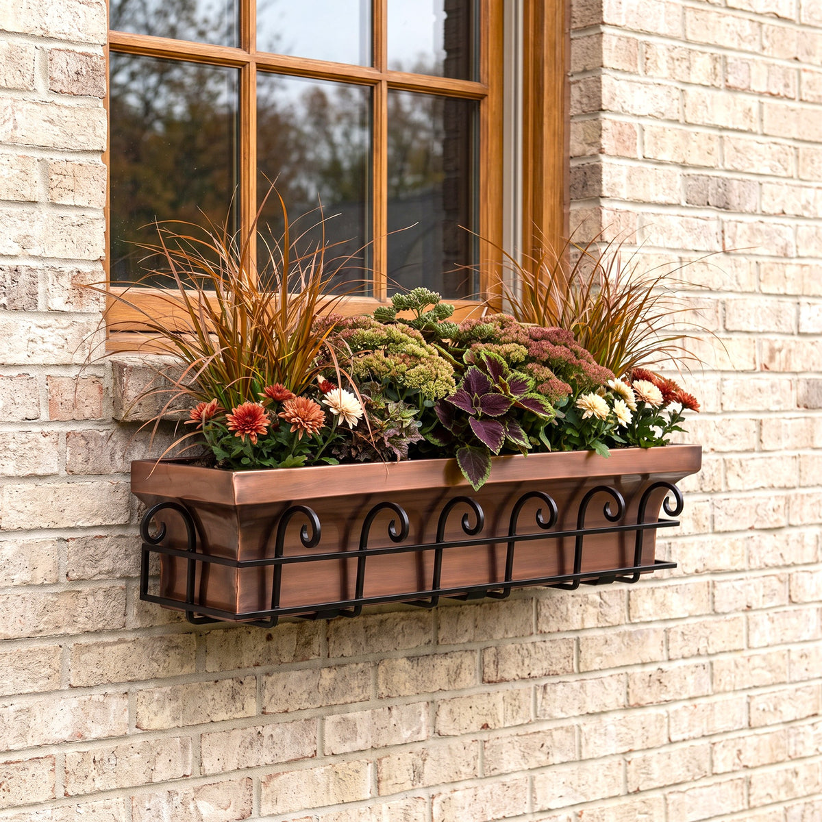 h potter brand copper-colored metal window box planter with a black scrollwork base, mounted on a beige brick wall beneath a wood-framed window and filled with ornamental grasses and flowers.
