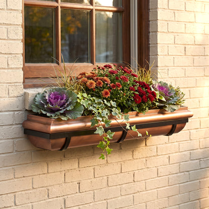 A copper-colored metal window box planter with black brackets, mounted on a beige brick wall beneath a wooden window and filled with colorful fall flowers and green foliage.