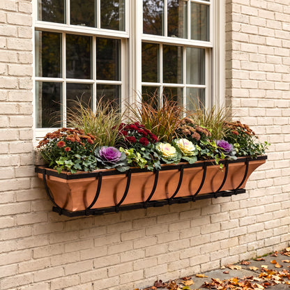 h potter brand copper window box planter with a black scrollwork frame, mounted on a beige brick wall beneath a white-framed window and filled with autumnal plants and flowers.