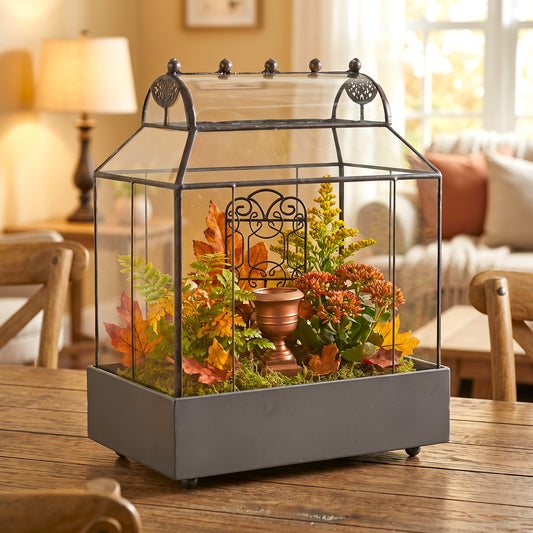 h potter brand tabletop terrarium with a clear glass panels, filled with faux autumn leaves, greenery, moss, and a small copper-toned centerpiece, set on a wooden table in a warm living room.