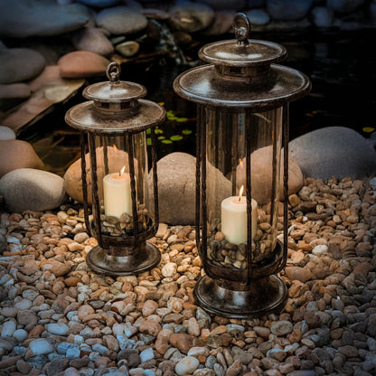 H Potter cast iron hurricane lanterns in large and small sizes with lit candles, displayed outdoors on river rocks for garden décor.