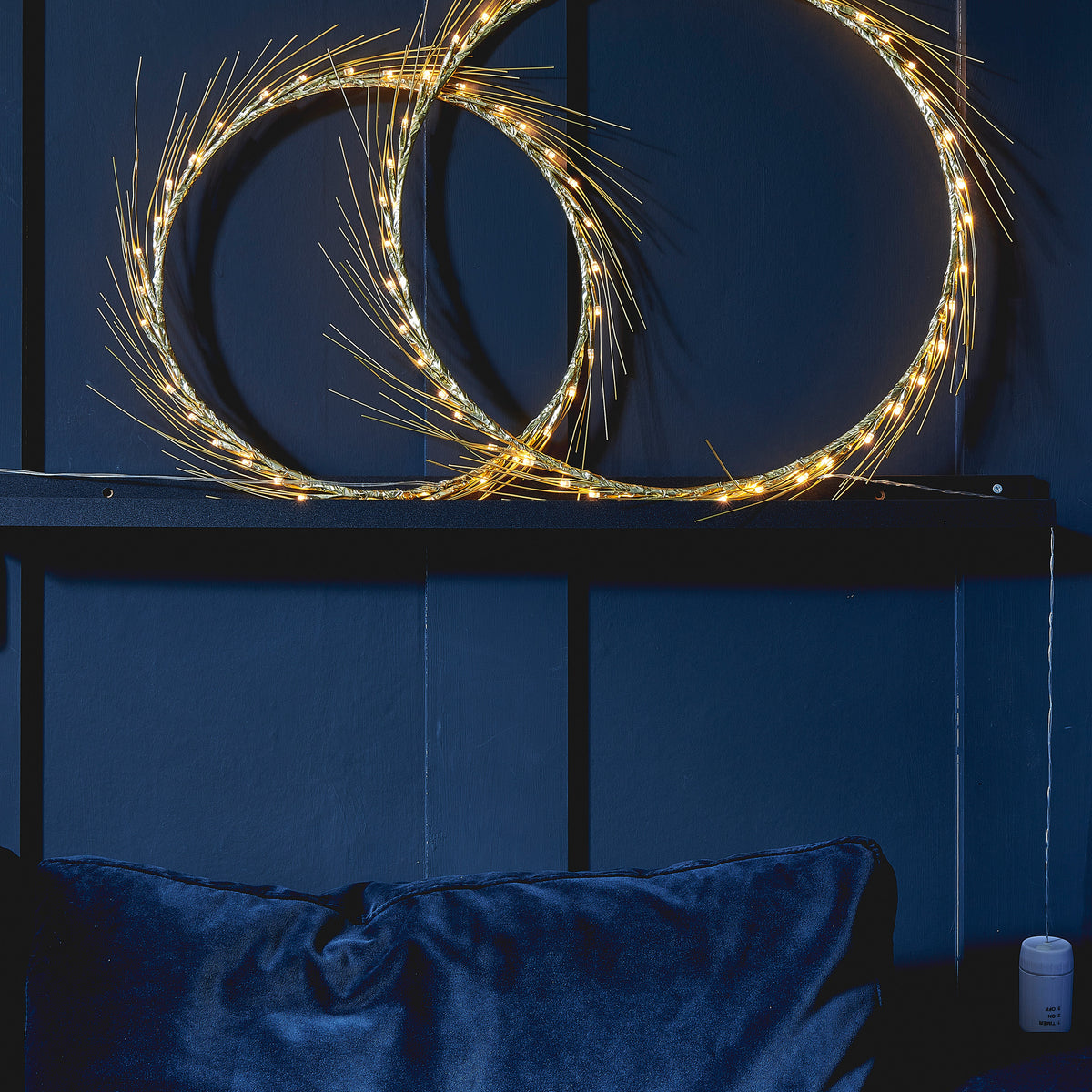 Large and small Celeste lighted wreaths resting on a dark shelf against navy panel