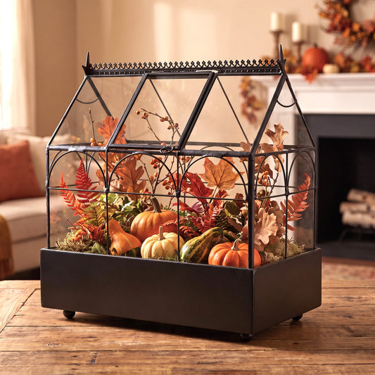 h potter brand terrarium planter with clear glass panels, filled with faux autumn foliage, small pumpkins, and gourds, displayed on a wooden table in a warm living room setting.