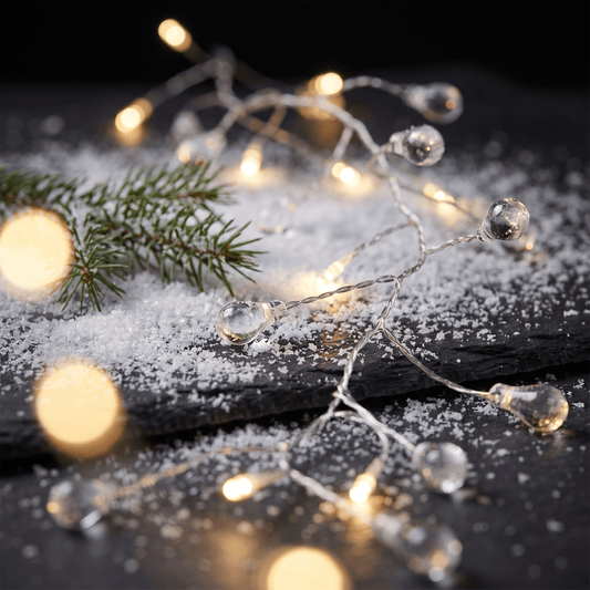 h potter LED string lights with clear teardrop-shaped glass bulbs emitting a warm yellow glow, draped over a dark slate surface with white faux snow, a pine branch, and soft lights.
