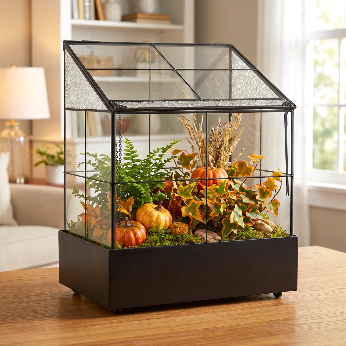 h potter brand house-shaped glass terrarium filled with autumn decorations including small pumpkins, ivy, ferns, and dried wheat, displayed on a wooden table in a bright living room.