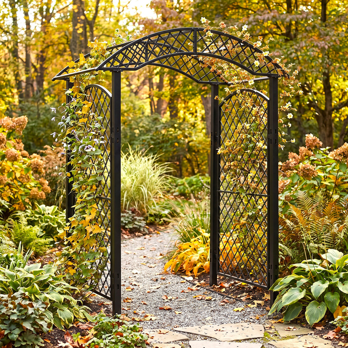 H Potter black metal garden arbor arch with lattice trellis panels stands open on a gravel path surrounded by autumn foliage and plants in a garden setting.