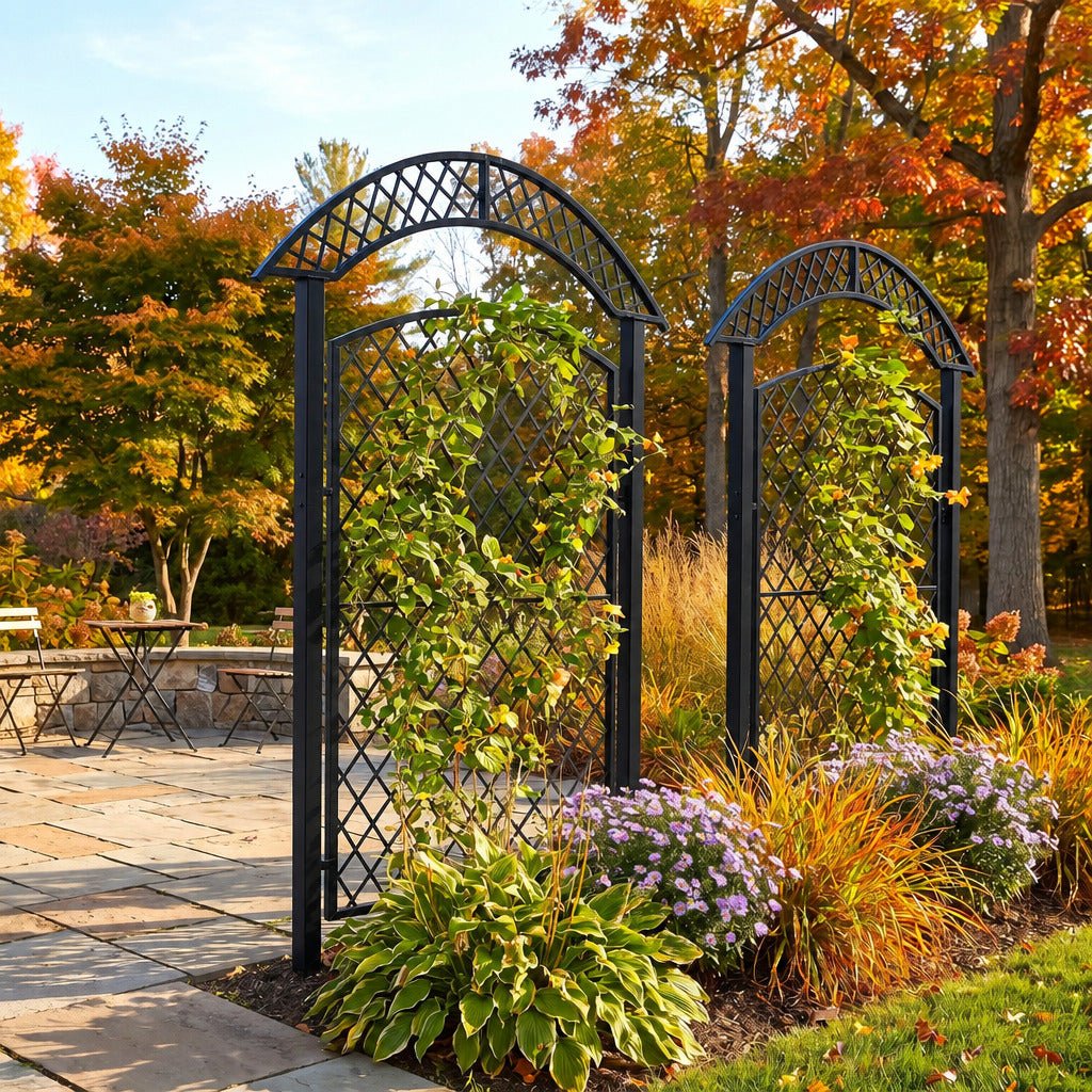 Two black metal arched trellis garden stakes with diamond lattice panels, shown in a landscaped outdoor garden bed with climbing green plants, purple flowers, ornamental grasses, and autumn trees in the background.