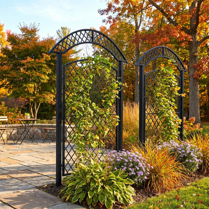 Two black metal arched trellis garden stakes with diamond lattice panels, shown in a landscaped outdoor garden bed with climbing green plants, purple flowers, ornamental grasses, and autumn trees in the background.