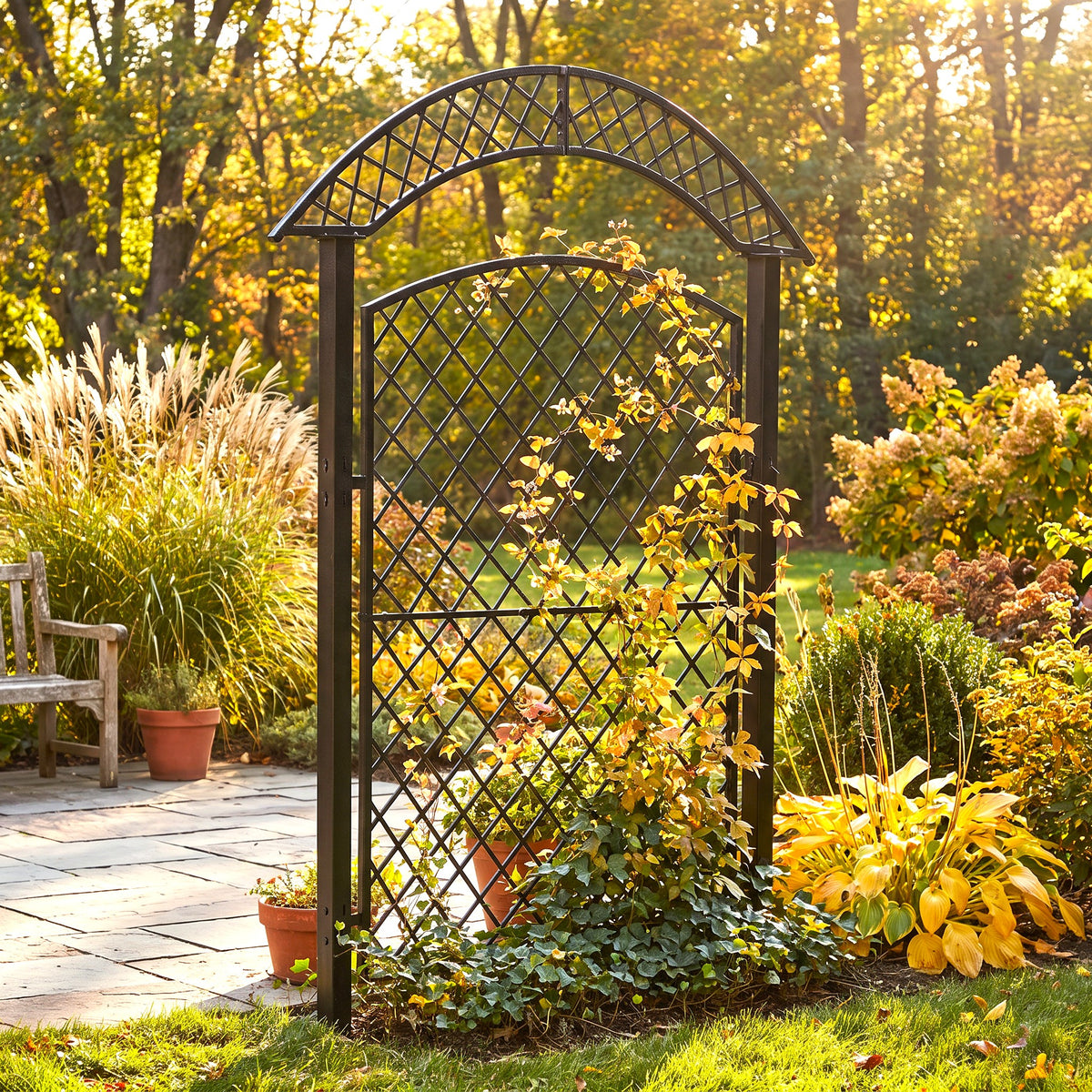H Potter black metal arched garden trellis with a lattice pattern, set in a sunny backyard garden with autumn foliage, potted plants, and a stone patio.