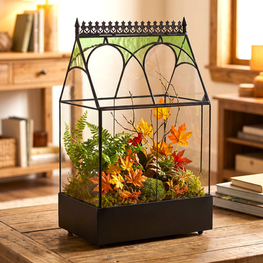 h potter brand terrarium with a house-shaped glass top and decorative finial, filled with faux moss, green ferns, and autumn-colored leaves, displayed on a wooden table in a warm, softly blurred room setting.