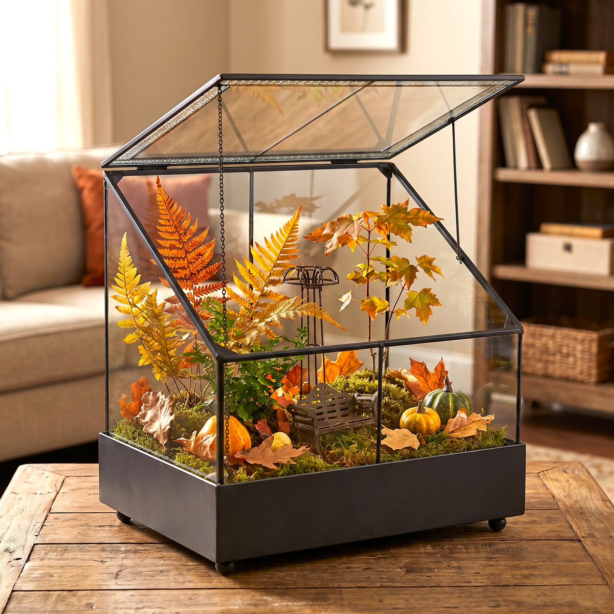 H Potter brand glass terrarium, filled with autumn foliage, faux moss, small pumpkins, displayed on a wooden table in a cozy living room setting.