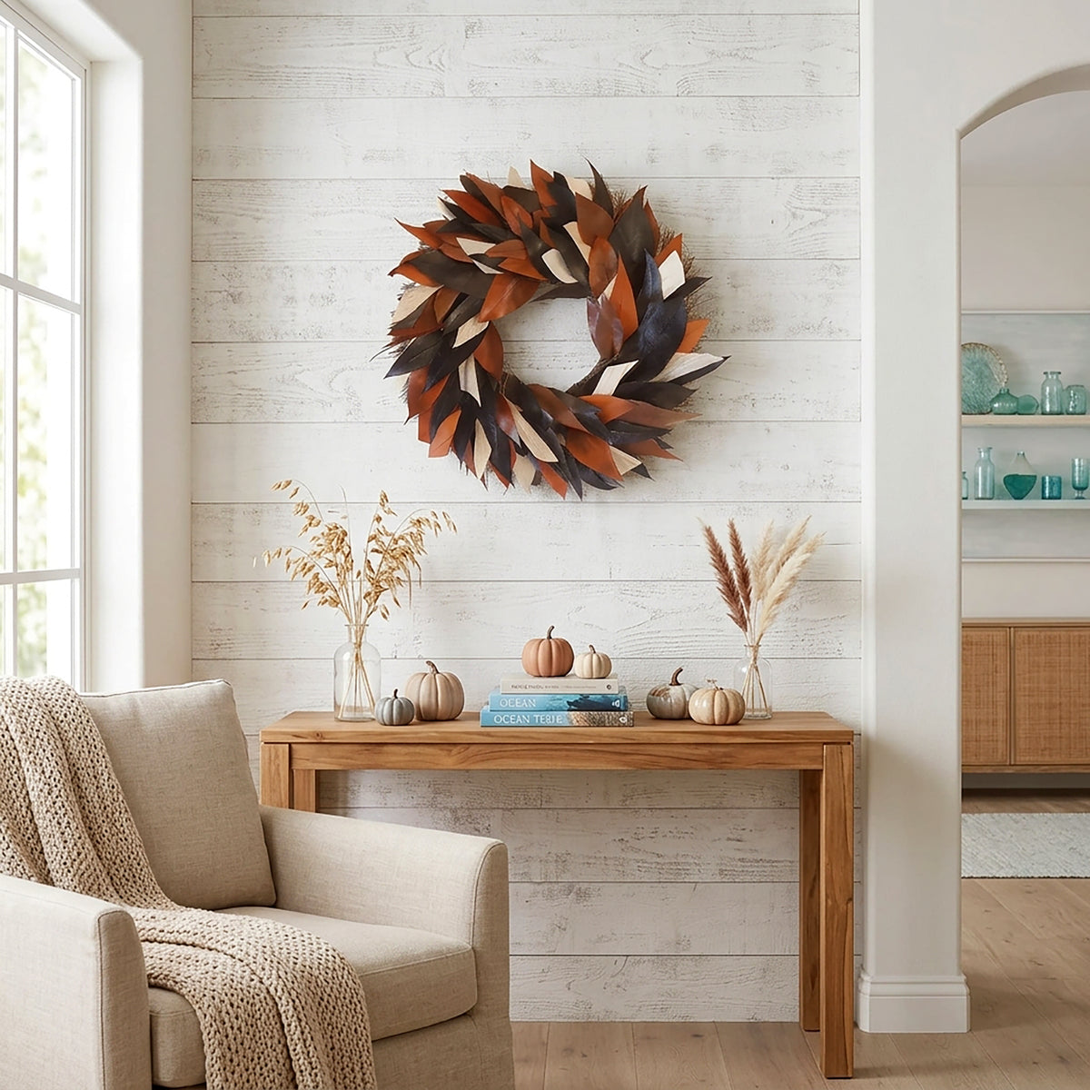 h potter brand decorative autumn leaf wreath in warm brown, rust, and cream tones hangs on a white shiplap wall above a wooden console table styled with pumpkins, books, and dried grasses in a bright living room.