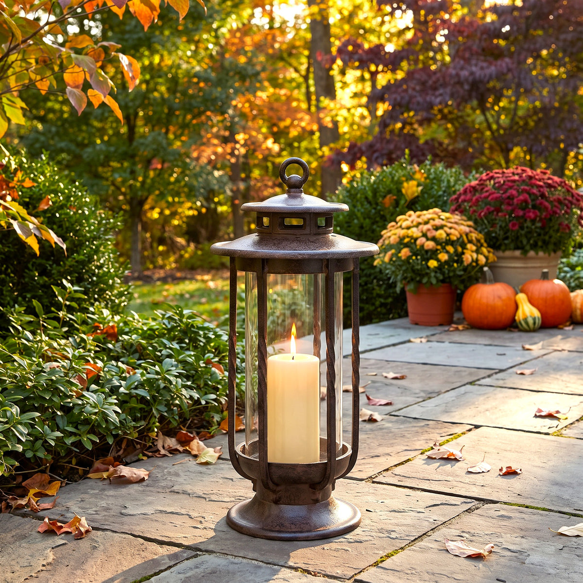 h potter cast iron lantern with a lit white pillar candle sits on a stone patio in an autumn garden setting with pumpkins and mums in the background.