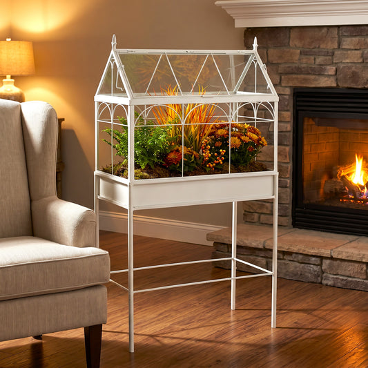 A white metal house-shaped garden planter with a glass roof and built-in tray, filled with greenery and autumn flowers, placed in a cozy living room next to an armchair and in front of a stone fireplace with a fire burning.