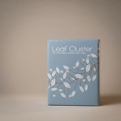 A rectangular product box for a 'Leaf Cluster' 150 LED battery operated light chain, featuring a light blue cover with white leaf illustrations and brown branch details, set against a neutral beige background.