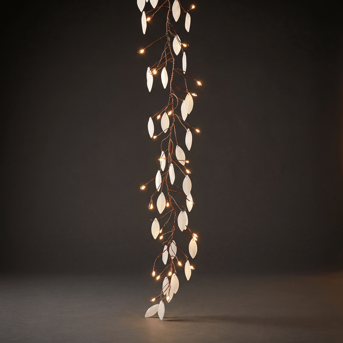 A vertical decorative string lights with white leaf-shaped lights and small warm white bulbs on a dark brown wire, photographed against a dark gray studio background.