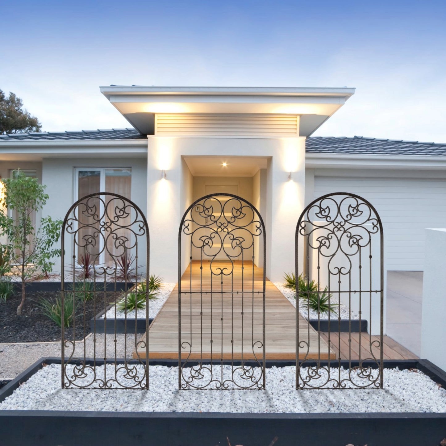 A set of three iron scrollwork garden trellises in front of a house entrance, with a well-lit exterior and greenery in the background.