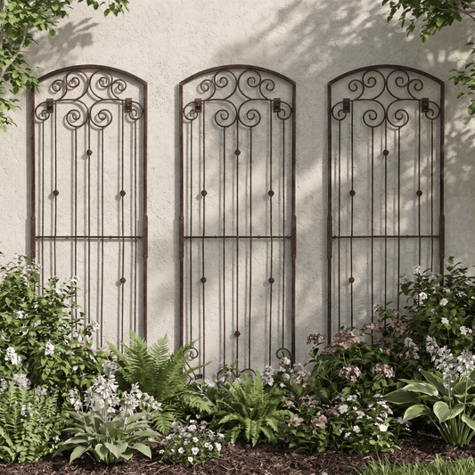 Three H Potter metal garden trellis with ornate scrollwork tops mounted on a stucco wall above a landscaped garden bed with green plants and small white flowers.