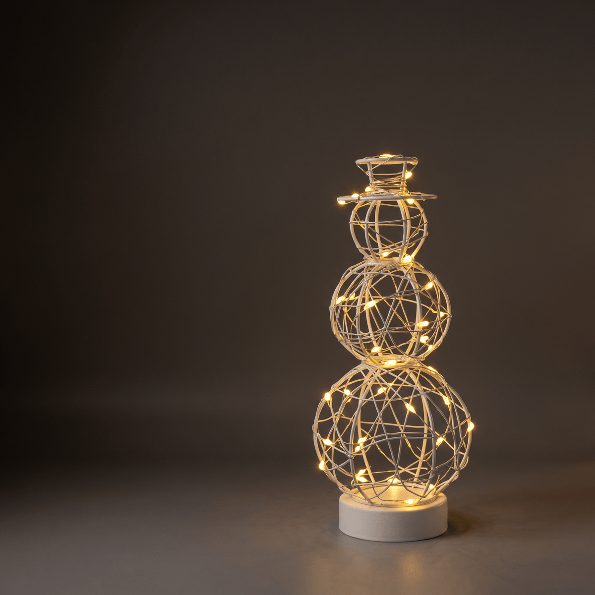 A snowman-shaped wire light sculpture with glowing warm white LED lights, standing on a round white base against a dark gray studio background.