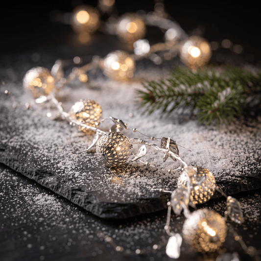 A close-up h potter orientale warm white string lights and textured gold ornaments, arranged on a dark surface with a dusting of white powder and a pine branch in the background.
