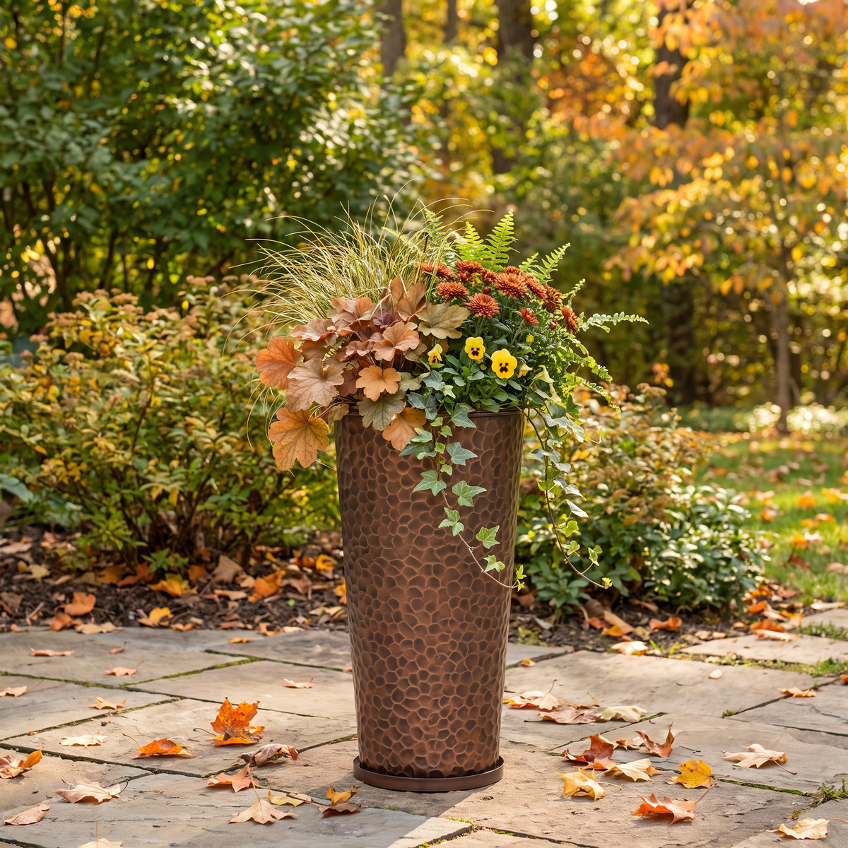 h potter brand tall, textured copper-colored metal planter filled with autumnal foliage and flowers, set on a stone patio in a garden with fallen leaves and colorful fall trees in the background.