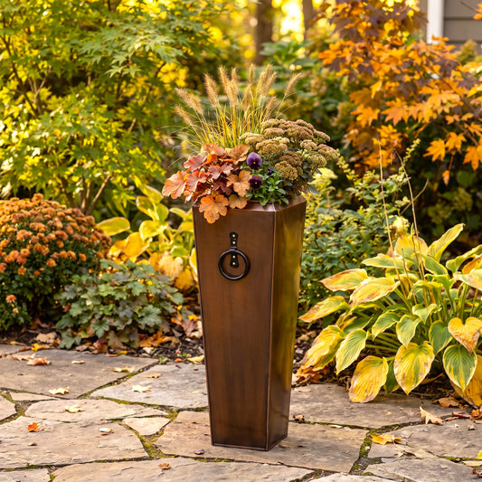 H Potter tall, narrow rectangular planter with a antique copper finish and a decorative black ring handle, filled with autumnal garden foliage, placed on a stone patio in a landscaped garden setting.