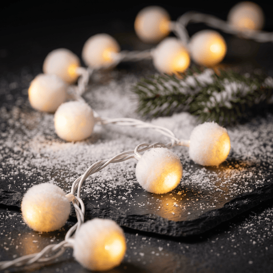 H Potter warm white LED ball string lights with fluffy pom-pom shades, arranged on a dark slate surface with scattered white faux snow and a green pine branch.