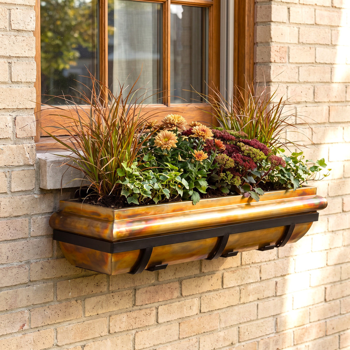 H Potter brand rectangular window box planter mounted on a beige brick wall beneath a wooden window, filled with ornamental grasses, green foliage, and mums in autumn colors.