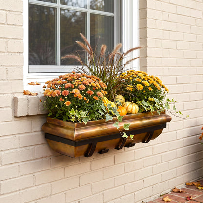H Potter brand wall-mounted rectangular planter box with a rustic copper finish, filled with orange and yellow mums, ornamental grass, ivy, and small pumpkins, installed on a beige brick wall beside a white-framed window.
