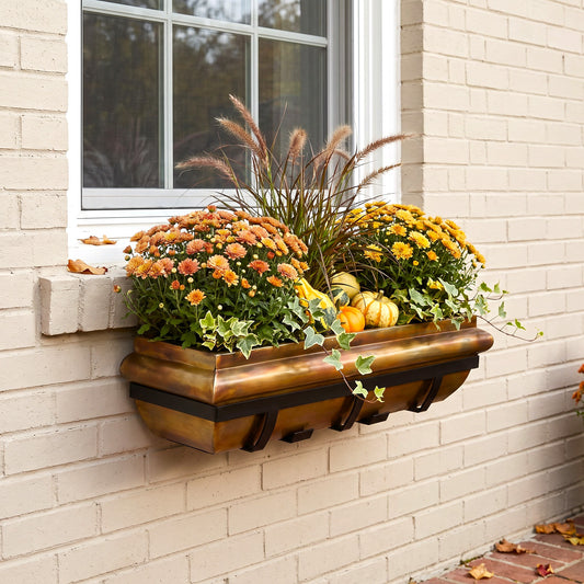 H Potter brand wall-mounted rectangular planter box with a rustic copper finish, filled with orange and yellow mums, ornamental grass, ivy, and small pumpkins, installed on a beige brick wall beside a white-framed window.