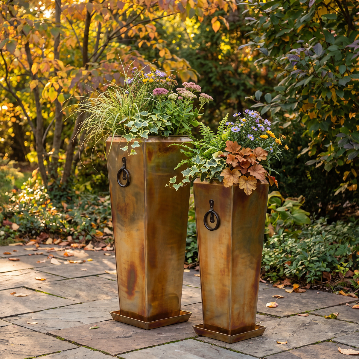 Two h potter brand tall rectangular metal planters with a distressed bronze-brown finish and black ring handles, filled with ornamental grass and autumn flowers, set on a stone patio in a garden with fall foliage.