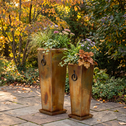 Two h potter brand tall rectangular metal planters with a distressed bronze-brown finish and black ring handles, filled with ornamental grass and autumn flowers, set on a stone patio in a garden with fall foliage.
